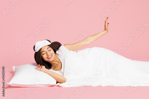 Calm young fun smiling woman wearing pyjamas jam sleep eye mask rest relax at home lay down on bed wake u pisolated on plain pastel light pink background studio portrait. Good mood night nap concept.