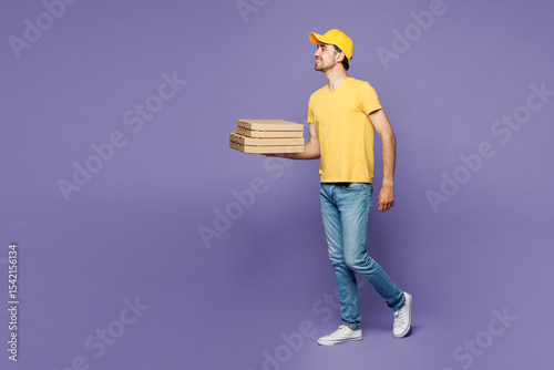 Full body young delivery guy employee man wearing yellow cap t-shirt uniform workwear work as dealer courier hold pizza in blank cardboard flatbox container isolated on plain pastel purple background.