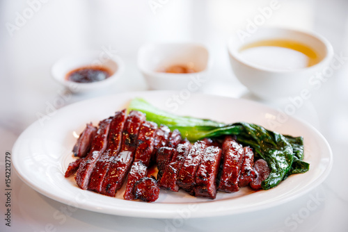 A plate of delicious Char Siu (Chinese BBQ pork) with steamed greens and dipping sauces, presented on a white background. A classic, savory Asian cuisine.