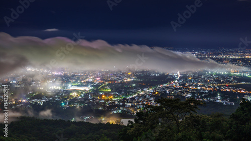 Aerial view of city skyline landscape at night with misty clouds over the illuminated cityscape from Doi Suthep mountain viewpoint in rainy season, Chiang Mai, Thailand.

