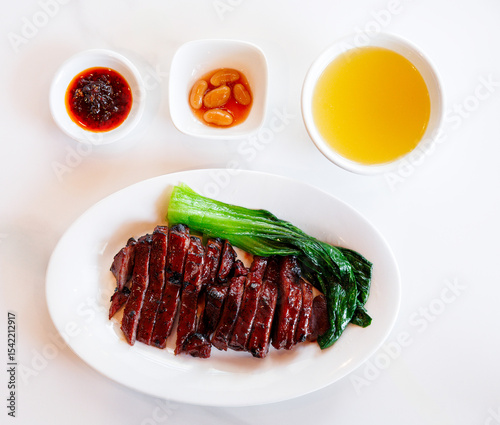 A plate of delicious Char Siu (Chinese BBQ pork) with steamed greens and dipping sauces, presented on a white background. A classic, savory Asian cuisine.