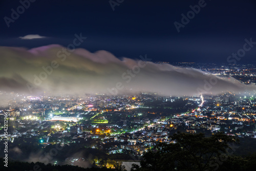 Aerial view of city skyline landscape at night with misty clouds over the illuminated cityscape from Doi Suthep mountain viewpoint in rainy season, Chiang Mai, Thailand.
