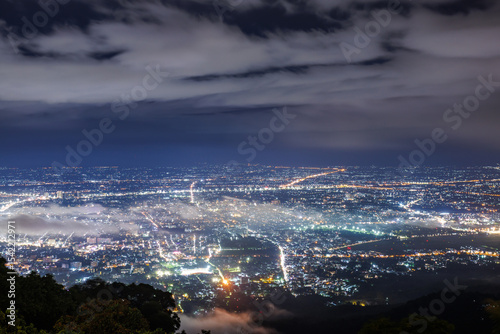 Aerial view of city skyline landscape at night with misty clouds over the illuminated cityscape from Doi Suthep mountain viewpoint in rainy season, Chiang Mai, Thailand.
