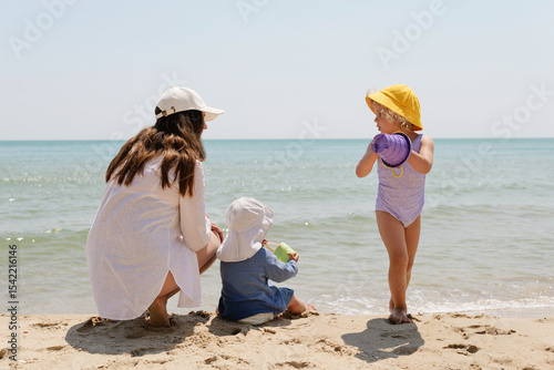 A mother and her two young children, facing the sea, enjoy a sunny day at the beach, with one child holding a purple bucket. Ideal for family summer concepts.