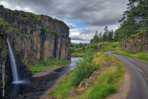Dramatic waterfall cascading into a valley, flanked by rugged cliffs and a winding road. Lush greenery surrounds the river and mountains