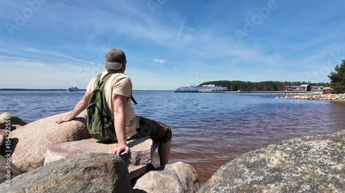 Contemplating the Serene and Picturesque Lakeside View on a Bright and Sunny Day. a traveler watches the passing cruise passenger liners, sitting on the rocks