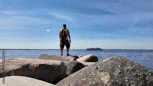 A solitary hiker is standing on a rocky shoreline while taking in the vast, tranquil waters with passing cruise ship