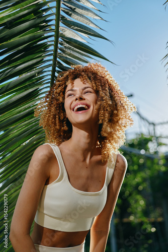 smiling woman with curly hair wearing white crop top outdoors under sunny sky by tropical palm leaves, joyful and relaxed, vibrant summer atmosphere