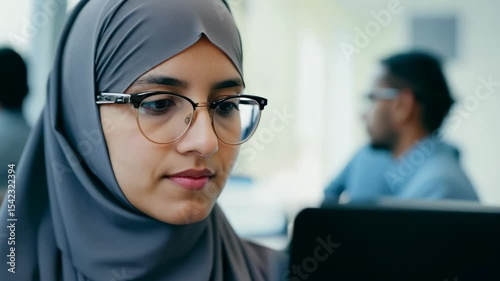 Young Caucasian woman in hijab with eyeglasses in front of laptop monitor in office. The young woman in hijab and eyeglasses expertly navigates her laptop.