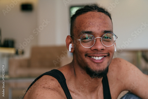Foto A headshot of an African American man with glasses and headphones, his face covered in severe acne, sitting on the living room floor
