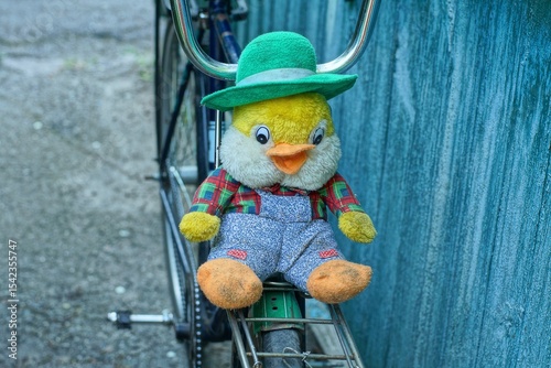 one toy soft plush baby in a green hat funny small with black eyes and an orange beak multi-colored in gray pants beautiful duckling lies on an iron bicycle rack near a wooden wall