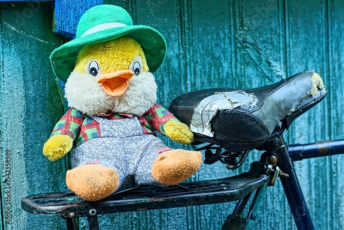 one toy soft plush baby in a green hat funny small with black eyes and an orange beak multi-colored in gray pants beautiful duckling lies on an iron bicycle rack and  saddle near a wooden wall
