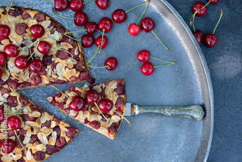 Close-up of a cherry pie from above, lying on a serving platter. A piece of cake has been cut off. There are fresh cherries next to the cake.