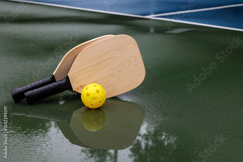 Two Pickleball paddles and one yellow ball next to a wet blue court in a neighborhood park
