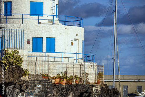 White and blue three-story lighthouse in the port on the Atlantic Ocean coast on the island of Tenerife against the background of a cloudy sky close-up	
 photo