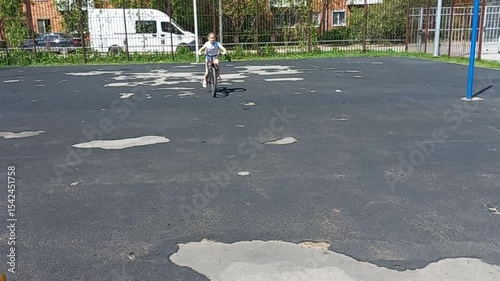 a nine year old girl rides a bicycle on cracked asphalt at a city playground, a boy with a ball runs across the road