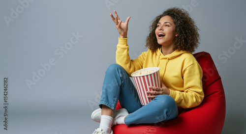 Cheerful young woman enjoying popcorn while sitting on a red bean bag chair, having fun and relaxing at home.