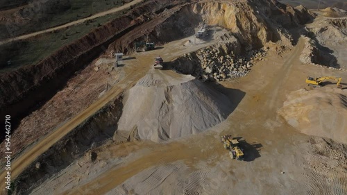 High aerial view of a wheel loader driving to the top of a massive gravel stockpile at a stone quarry, with other industrial machinery working in the background.
