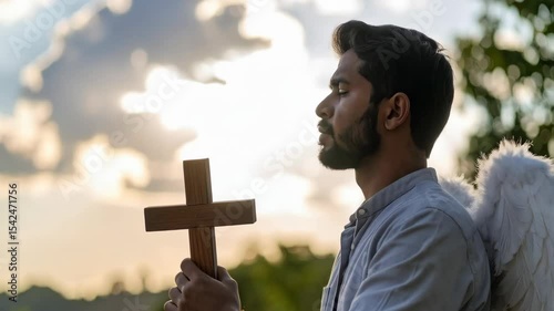 Bearded man dressed as angel holding wooden cross in contemplative prayer against sunlit sky and green foliage