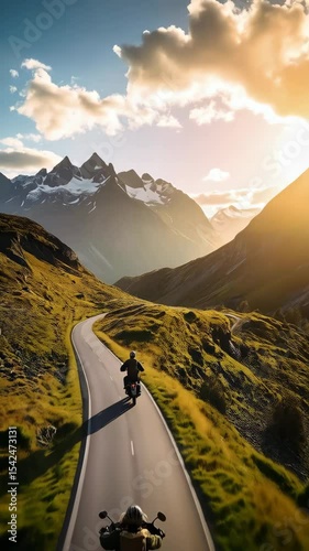 Motorcycle riders traveling on a scenic mountain road during golden hour with snow capped peaks and beautiful cloud formations