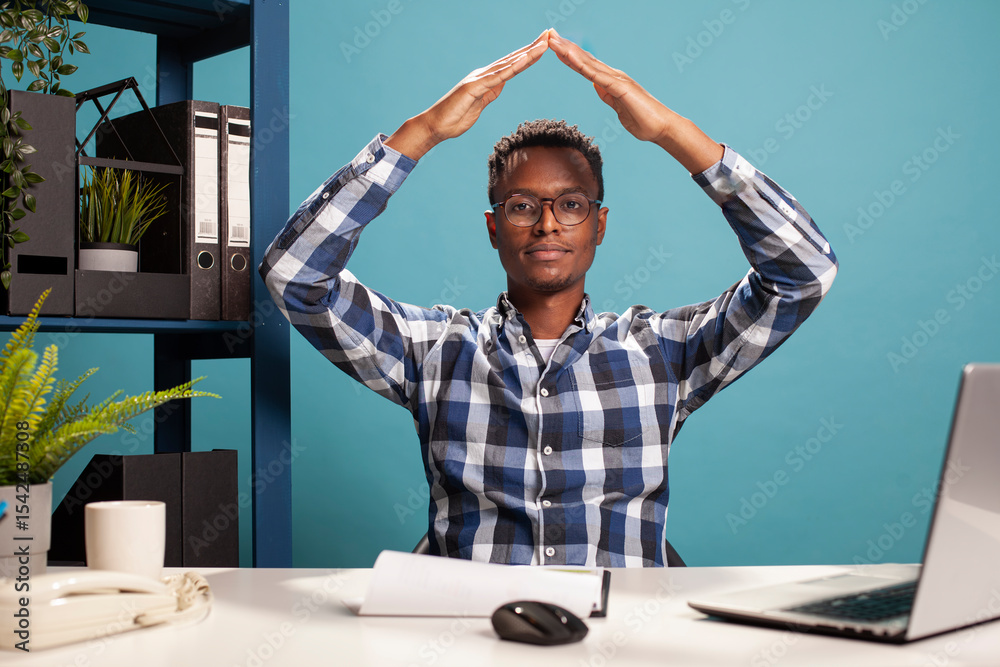 Fototapeta premium African American man wearing glasses, seated at office desk, making a triangle with his arms above head. Male analyst forming a safety roof gesture with his hands, symbolizing confidence and security.