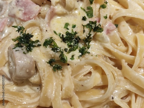 A close-up image of creamy pasta, likely fettuccine Alfredo, topped with chopped parsley. The dish includes mushrooms, bits of ham or bacon, and is coated in a rich white sauce with grated cheese.