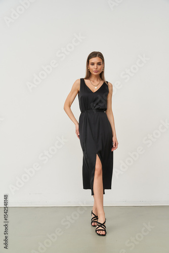 Woman in elegant sleeveless black dress with side slit, standing confidently, hands relaxed, wearing black sandals, on neutral floor. She has straight hair and a subtle smile