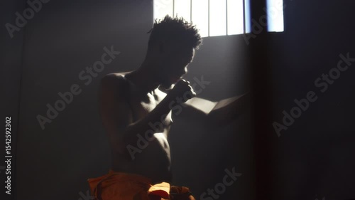 Shirtless Black male inmate practicing shadowboxing during workout in prison cell with light streaming through barred window