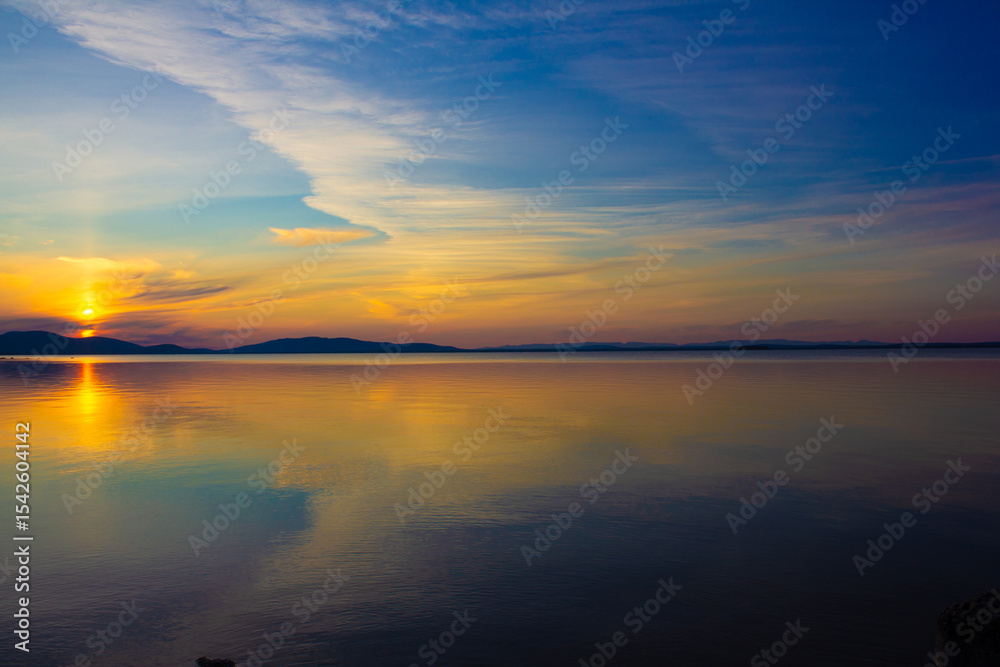 Obraz premium Stunning sunset over calm lake with vibrant colorful sky, dramatic clouds, and perfect reflection in water. Serene nature landscape ideal for backgrounds, banners, travel, meditation, and relaxation.