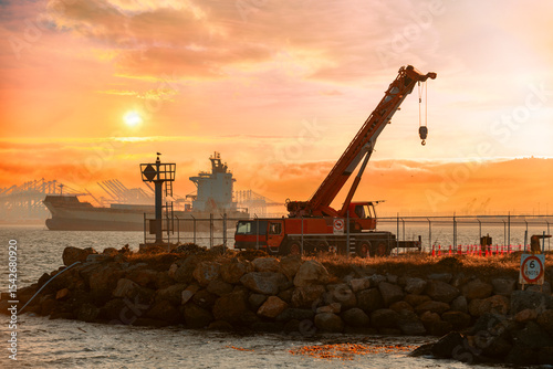 On a rocky shoreline, a mobile crane stands silhouetted against a vibrant sunset.