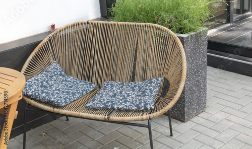 An outdoor wicker chair with navy blue floral cushions, next to a grey stone plant pot on the street