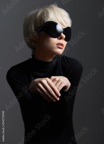 Stylish woman in black turtleneck and oversized sunglasses, posing delicately with hands, evoking a sense of high fashion and modern elegance against a dark backdrop.