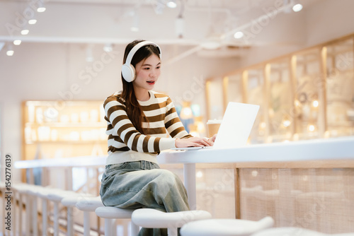 A young woman wearing headphones and glasses, appearing to be an East Asian female, intently uses a white laptop at a cafe table. photo
