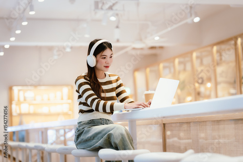 A young woman wearing headphones and glasses, appearing to be an East Asian female, intently uses a white laptop at a cafe table. photo