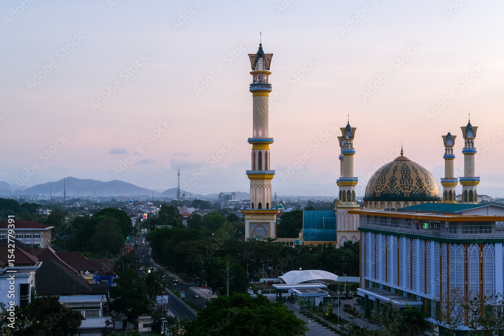 Fototapeta premium Golden twilight blankets the majestic mosque in Mataram city, Lombok, highlighting elegant minarets, vibrant streets, and surrounding hills under a serene pastel sky. Masjid Islamic Center Mataram.
