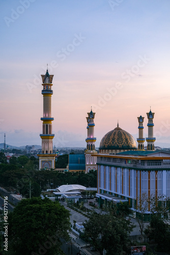 Golden twilight blankets the majestic mosque in Mataram city, Lombok, highlighting elegant minarets, vibrant streets, and surrounding hills under a serene pastel sky. Masjid Islamic Center Mataram.