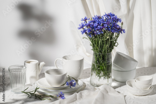 Bouquet of cornflowers in glass vase on table with white tableware. Summer still life.