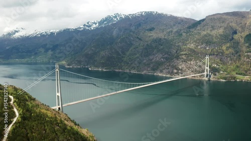 Hardanger Bridge gracefully spans vast body of fjord with towering mountains forming dramatic backdrop. Modern suspension bridge with prominent towers