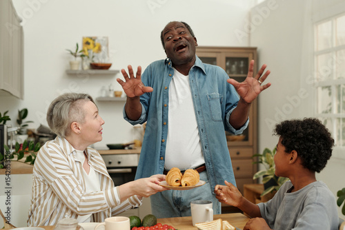 Family having breakfast together in modern kitchen, sharing food and laughter. Man appearing to sing or talk humorously while interacting with others at table