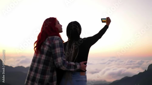 Couple embrace taking selfie atop mountain range overlooking cloudscape during sunset