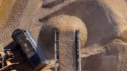 Top-down aerial of a conveyor belt dropping crushed stone onto a pile. A wheel loader works at the quarry, producing construction materials like sand and gravel.
