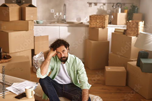 A man sits on the floor surrounded by numerous boxes as he takes a moment to think while preparing for the move to his new home, reflecting on the changes ahead.