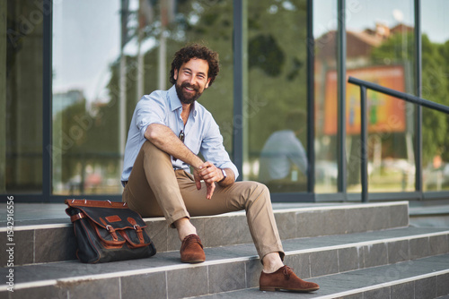 A smiling man dressed in smart casual clothing sits comfortably on outdoor stairs, taking a break with a messenger bag beside him in an urban setting.