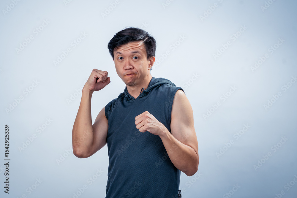 Naklejka premium Asian man in sleeveless hoodie showing excited expression while flexing arms and clenching fists, posing cheerfully with energy and confidence in front of plain light studio background