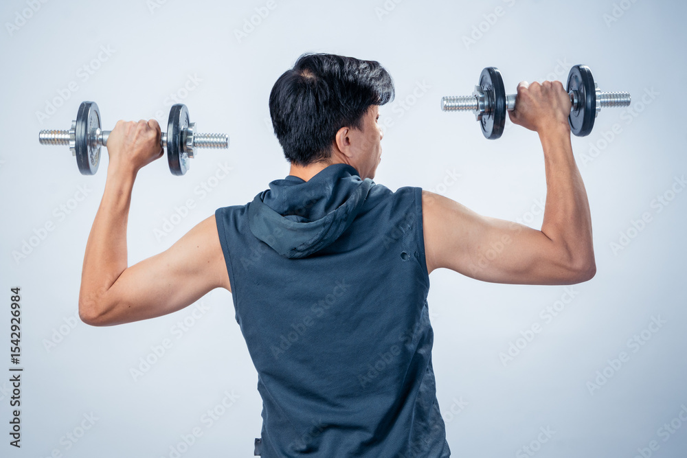 Asian man in sleeveless hoodie lifting dumbbells at shoulder height with both arms from the back, showing muscular build, workout posture, and fitness routine in studio with plain background Asian man in sleeveless hoodie lifting dumbbells at shoulder height with both arms from the back, showing muscular build, workout posture, and fitness routine in studio with plain background