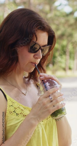 Elegant woman in stylish sunglasses drinks refreshing beverage and relaxes in city park. brunette female enjoys flavor of drink and sights of park. Vertical shot