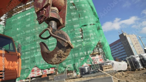 Steel crane hook hangs suspended above bustling construction zone. Sunlight glints off polished surface as workers prepare rigging below