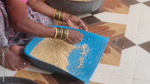 A woman housewife, is winnowing jowar (sorghum) grains outside her rural home. Separating the grains from the chaff using a traditional flat basket, an essential process to clean edible grains.