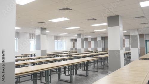 Neatly folded cafeteria tables stand piled high in empty school dining hall after lunchtime. Metallic legs gleam under fluorescent lights