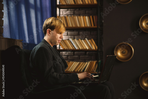 Man working on laptop in a dark, atmospheric room. Sitting in a chair, focused, with bookshelves and curtains in the background. Business, education, remote work concept.